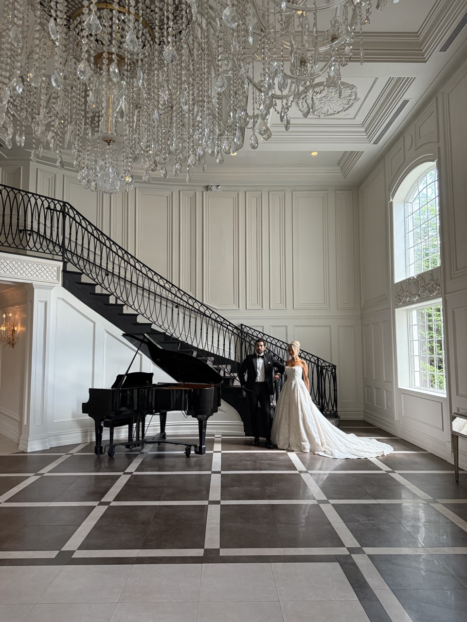 Bride and groom stand beneath a sweeping staircase and crystal chandelier in the grand foyer of Florentine Gardens in River Vale, New Jersey, with a black grand piano and elegant architectural details creating a timeless wedding portrait.